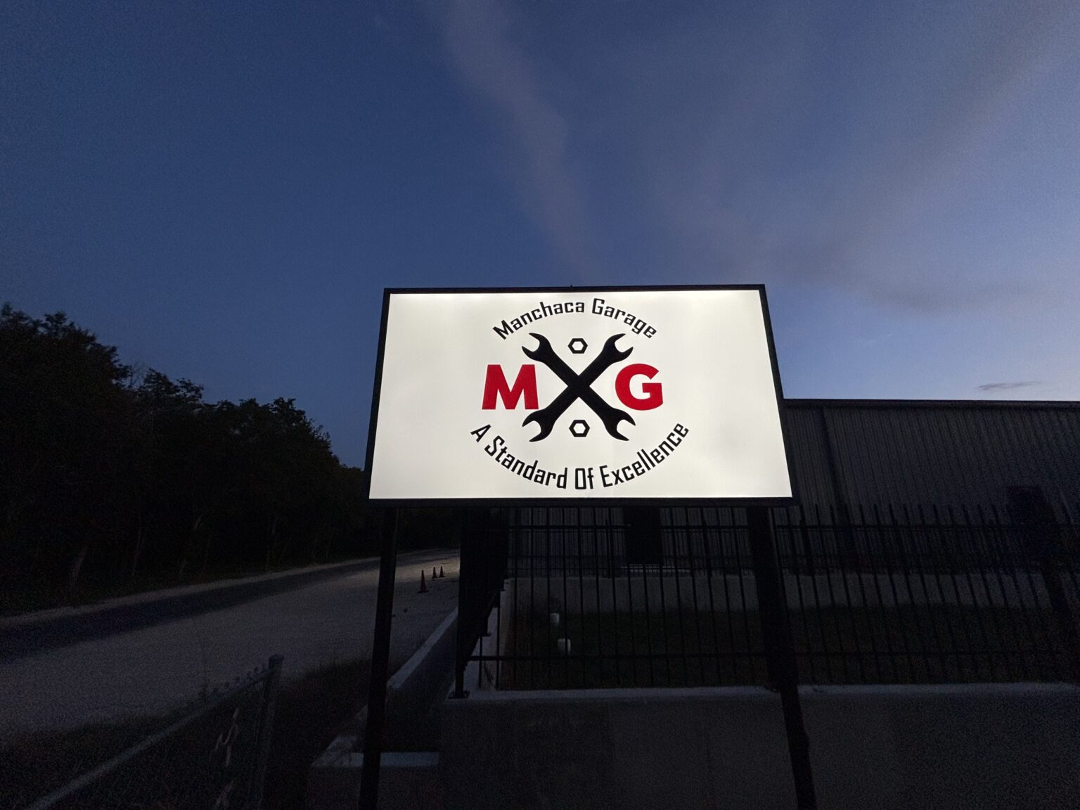 The illuminated Manchaca Garage sign with the MG crossed-wrench logo outside the shop